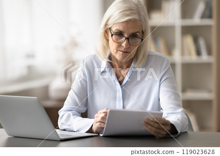 Older woman reading document seated at desk with laptop Older woman reading document seated at desk with laptop 119023628
