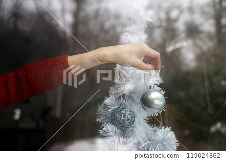 Female hand in festive red sweater placing shiny blue holiday bauble on white christmas tree 119024862