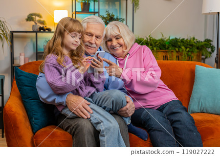 Child girl granddaughter teaching grandfather and grandmother using smartphone on sofa in home room Child girl granddaughter teaching grandfather and grandmother using smartphone on sofa in home room 119027222
