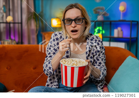 Excited amazed woman sitting on couch eating popcorn and watching interesting TV serial film game Excited amazed woman sitting on couch eating popcorn and watching interesting TV serial film game 119027301