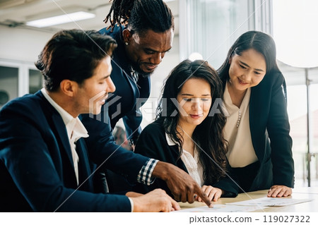 International business professionals gather in a meeting room, led by a female boss. Their teamwork is evident as they discuss financial results, share ideas, and brainstorm solutions for success. 119027322