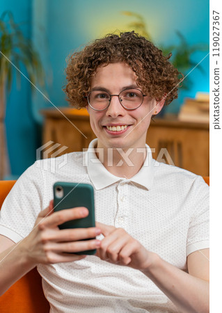 Caucasian young man sitting on couch using mobile smartphone share messages smiles at home room Caucasian young man sitting on couch using mobile smartphone share messages smiles at home room 119027367