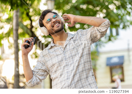 Happy Indian man in wireless headphones choosing, listening disco music on smartphone on street 119027394