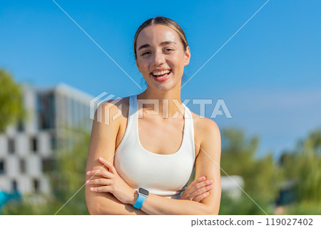 Portrait of happy Caucasian young sporty woman in activewear standing arms crossed in city park 119027402