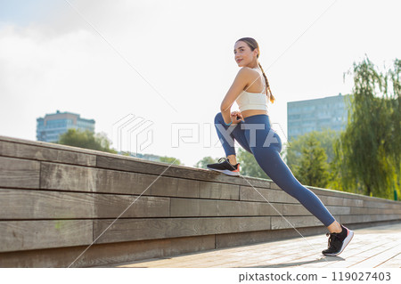 Athletic young woman runner stretching legs before jogging on bridge in city park on sunny day 119027403