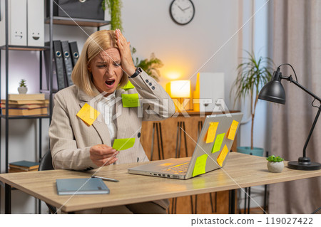 Exhausted Caucasian mature businesswoman with sticky notes and laptop sitting at home office desk 119027422