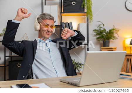 Happy young businessman wearing sunglasses and wireless headphones enjoy music at home office desk Happy young businessman wearing sunglasses and wireless headphones enjoy music at home office desk 119027435