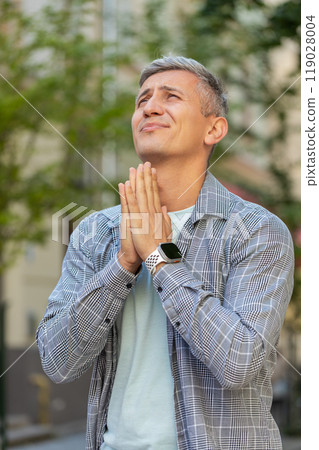 Caucasian mature man tourist praying with closed eyes to God asking for blessing on city town street 119028004