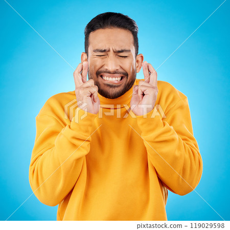 Nervous, eyes closed and fingers crossed by asian man with anxiety in studio for news, feedback or review on blue background. Hand, emoji and guy with stress for results, competition or giveaway 119029598