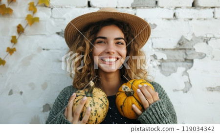 Smiling Young Woman in Straw Hat Holding Two Pumpkins Against White Wall, Autumn Harvest Concept 119034342