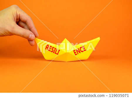 Resilience symbol. Concept word Resilience on beautiful yellow paper boat. Beautiful orange background. Businessman hand. Business resilience concept. Copy space. Resilience symbol. Concept word Resilience on beautiful yellow paper boat. Beautiful orange background. Businessman hand. Business resilience concept. Copy space. 119035071