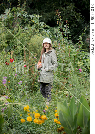 Girl wearing a jacket and a white hat walks through the garden with flowers holding a pumpkin 119035818