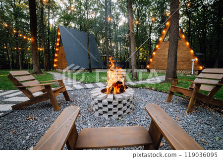 Campfire area with wooden chairs and cabins in background, creating a cozy outdoor gathering space Campfire area with wooden chairs and cabins in background, creating a cozy outdoor gathering space 119036095