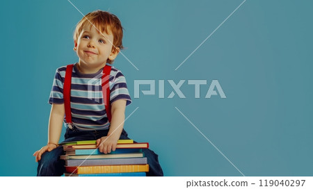 Cheerful child in striped shirt sitting on stack of colorful books against blue background 119040297