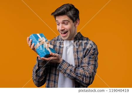 Handsome man holding gift box on yellow studio background and smiles to camera. Happy european guy received present and interested in what's inside. 119040419