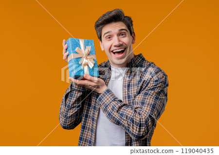 Handsome man holding gift box on yellow studio background and smiles to camera. Happy european guy received present and interested in what's inside. 119040435