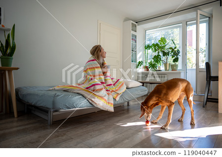 Happy woman dog owner enjoys slow morning coffee in bed wrapped in blanket dreamful looks at window. 119040447