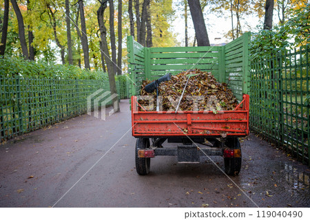 Small dump truck with dry fallen leaves in autumn park riding on asphalt road. 119040490