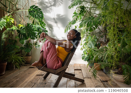Playful happy black woman sit on chair surround by tropical houseplants look at camera enjoy moment. Playful happy black woman sit on chair surround by tropical houseplants look at camera enjoy moment. 119040508
