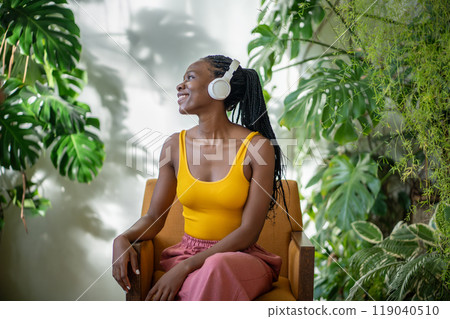 Happy black woman relaxing in armchair in cozy home with monstera plant, listen music in headphones Happy black woman relaxing in armchair in cozy home with monstera plant, listen music in headphones 119040510