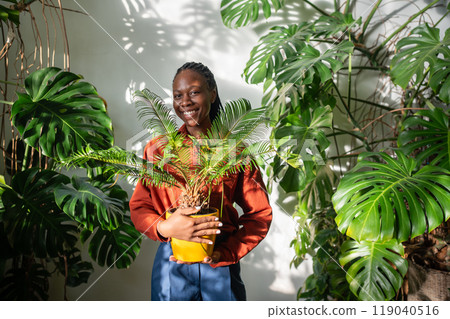 Cheerful pleased black woman plant lover holding Cycas plant in yellow pot in hands look at camera 119040516