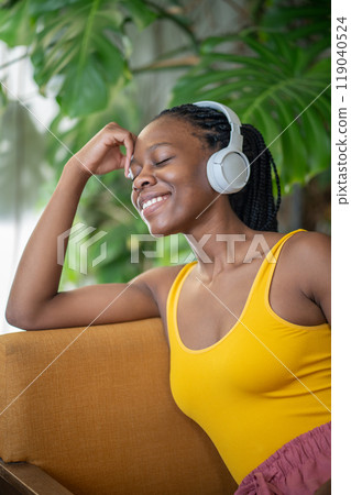 Carefree black woman relaxing in armchair at cozy home with houseplant, listen music in headphones Carefree black woman relaxing in armchair at cozy home with houseplant, listen music in headphones 119040524