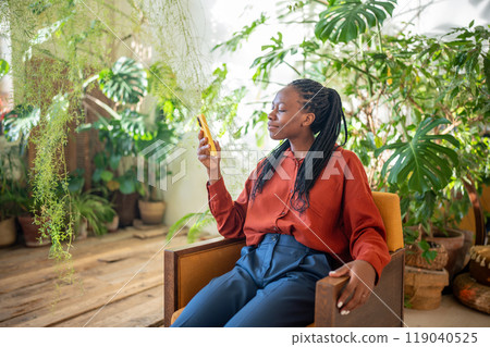 Relaxed pleased african american woman sit on armchair with smartphone surround by tropical plants. 119040525