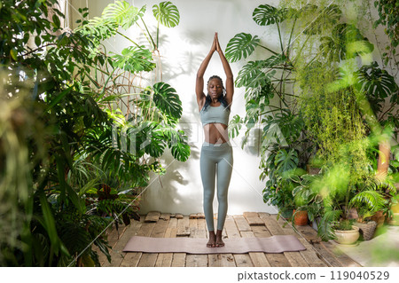 Serene balanced black woman stands on yoga mat surround by plants meditating feel calmness, harmony Serene balanced black woman stands on yoga mat surround by plants meditating feel calmness, harmony 119040529