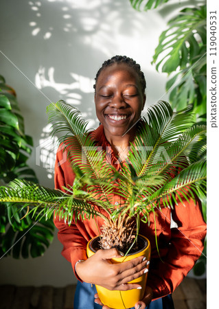 Happy black woman hugging pot with Cycas plant. Joyful smiling African American girl at flower shop 119040531
