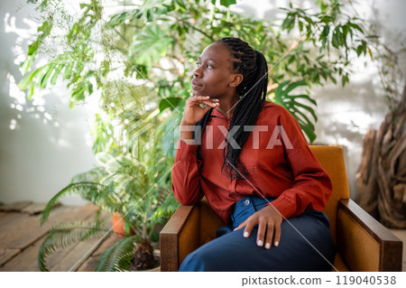 Thoughtful african american woman sitting on armchair on houseplants background pondering look aside Thoughtful african american woman sitting on armchair on houseplants background pondering look aside 119040538