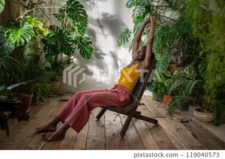 Relaxed african american young woman stretching arms up smiling sits on chair in urban jungle room. Relaxed african american young woman stretching arms up smiling sits on chair in urban jungle room. 119040573