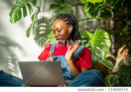 Interested black woman in earphones with laptop make video call relaxed sits on monstera background. 119040574