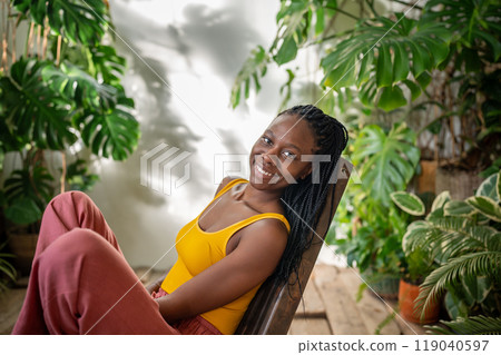 Pleased relaxed black woman looking at camera rest on cozy chair surround by tropical indoor plants. 119040597