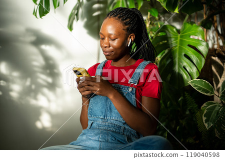 Relaxed black girl in earphones holds smartphone typing message, listen music in room with monstera. 119040598