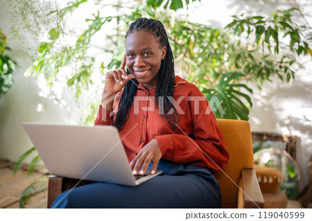 Happy pleased black woman with laptop on laps sitting on chair in sunny room smiling look at camera. Happy pleased black woman with laptop on laps sitting on chair in sunny room smiling look at camera. 119040599