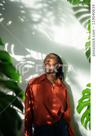 Happy pleased african american young woman standing leaning wall enjoying sunbeams with closed eyes. 119040634