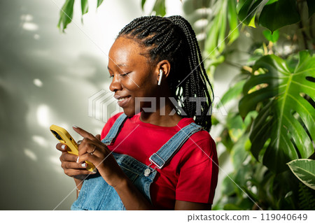 Relaxed black woman in earphones using smartphone at home, selects favourite music track in app. 119040649