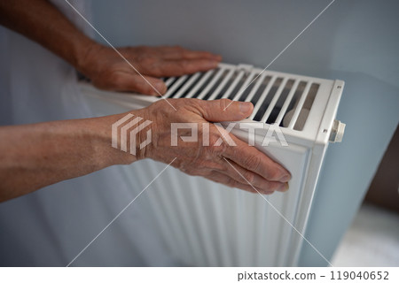 Mature woman hands touching heater checking temperature in heating season in cold weather at home. 119040652