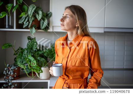 Smiling relaxed woman plant lover in orange dress holding mug in cozy kitchen, look at camera Smiling relaxed woman plant lover in orange dress holding mug in cozy kitchen, look at camera 119040678