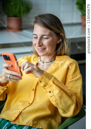 Happy Swedish woman with smartphone chatting with friends sitting in chair in kitchen and relaxing. Happy Swedish woman with smartphone chatting with friends sitting in chair in kitchen and relaxing. 119040679