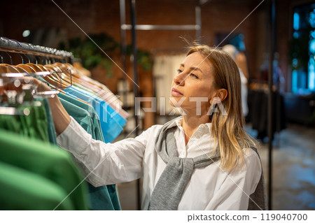 Delighted fashionable woman examines and chooses garments on racks in clothing store. Sorting shirts 119040700