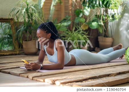 Relaxed black woman lying on fitness mat holding smartphone take break after home sported training. 119040704