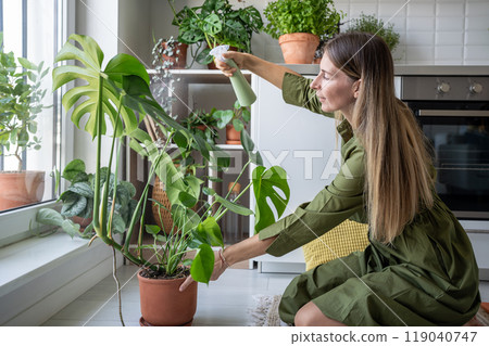Pleased satisfied woman plant lover taking care about green houseplants spraying water to monstera. 119040747