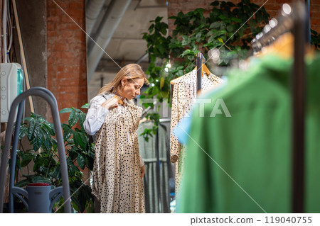 Fashionable woman contentedly picks garments and tries dress from hangers by mirror in shopping mall 119040755