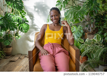 Happy African woman relaxing on chair in cozy apartment with houseplants, black girl resting at home Happy African woman relaxing on chair in cozy apartment with houseplants, black girl resting at home 119040757