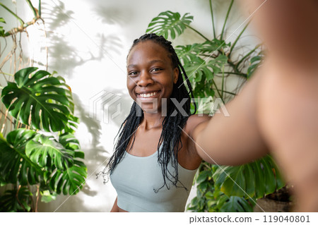 Happy african american woman smiling making selfie on smartphone surround by tropical houseplants. 119040801