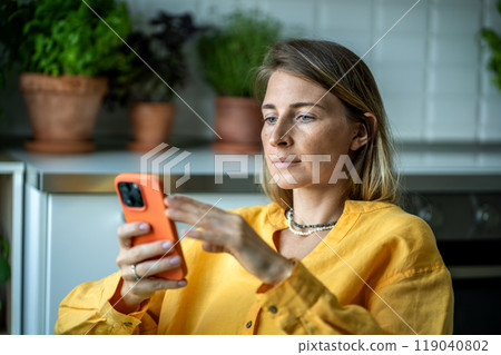 Thoughtful relaxed European woman looking to screen cellphone reading message, scrolling web at home 119040802