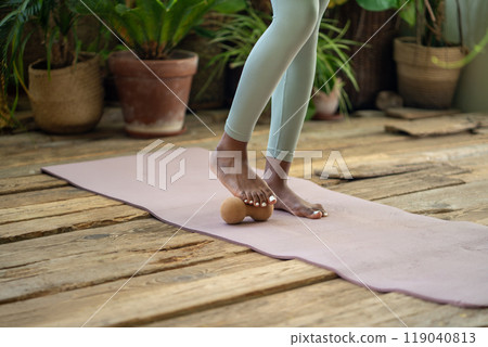 Black woman stands on gym mat using massage ball to roll out tension feet spend time for self-care. Black woman stands on gym mat using massage ball to roll out tension feet spend time for self-care. 119040813
