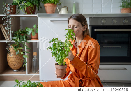Woman enjoying smell of basil. Girl sits on floor, inhales aroma of home-grown flavour basil in pot Woman enjoying smell of basil. Girl sits on floor, inhales aroma of home-grown flavour basil in pot 119040868