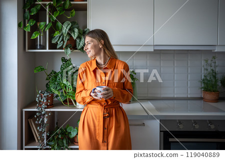 Smiling carefree woman drinking tea in kitchen among houseplants, looking out window. Slow life. Smiling carefree woman drinking tea in kitchen among houseplants, looking out window. Slow life. 119040889